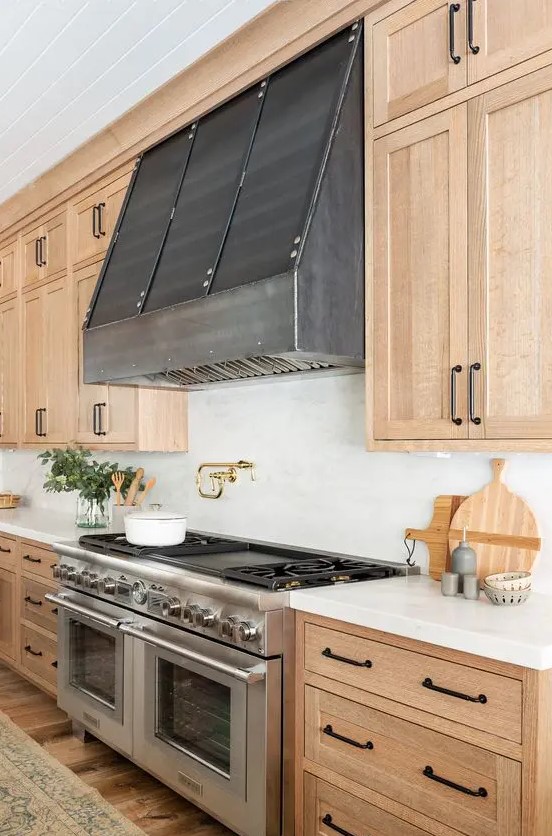 A chic light wood kitchen with shaker style cabinets, white stone countertops and backsplash, metal hood and black handles