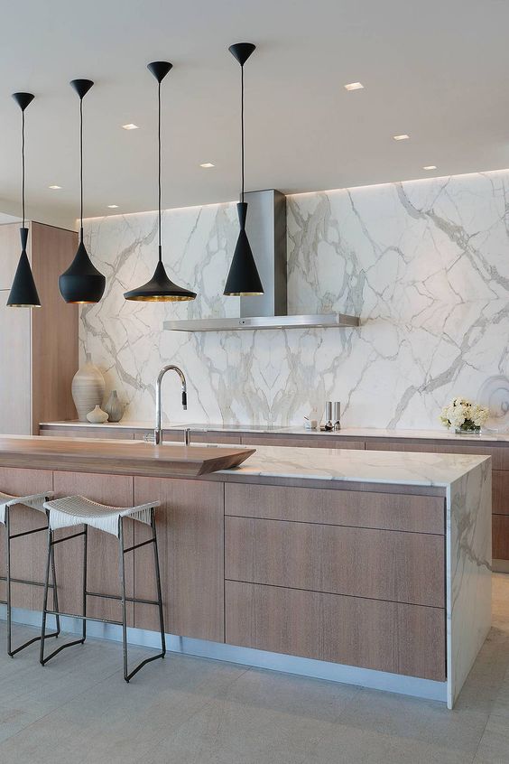 a minimalist kitchen with light stained cabinets and a white marble wall and countertop, a set of black and gold pendant lamps