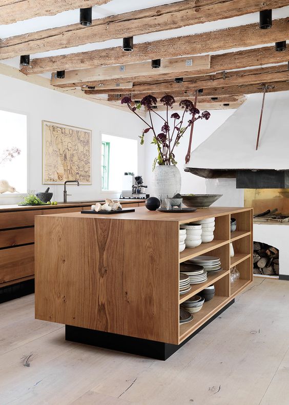 a modern kitchen with stained sleek cabinets, a matching kitchen island with open shelving and rough wooden beams and a fireplace