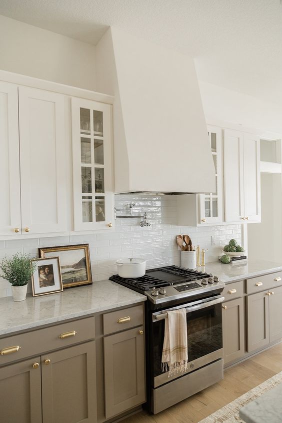 A delicate kitchen with white upper cabinets and taupe lower cabinets, a white tile backsplash and gold knobs for a chic and elegant touch