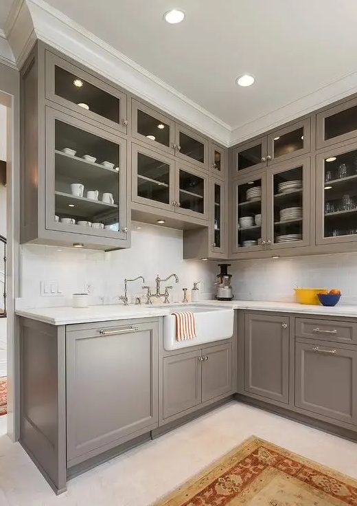 A sleek taupe vintage kitchen with shaker-style cabinets, glass tops, a white stone countertop and backsplash, and vintage faucets and knobs is all cool