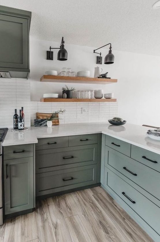 A stylish sage green kitchen with shaker style cabinets, white countertops and a white thin tile backsplash and floating shelves