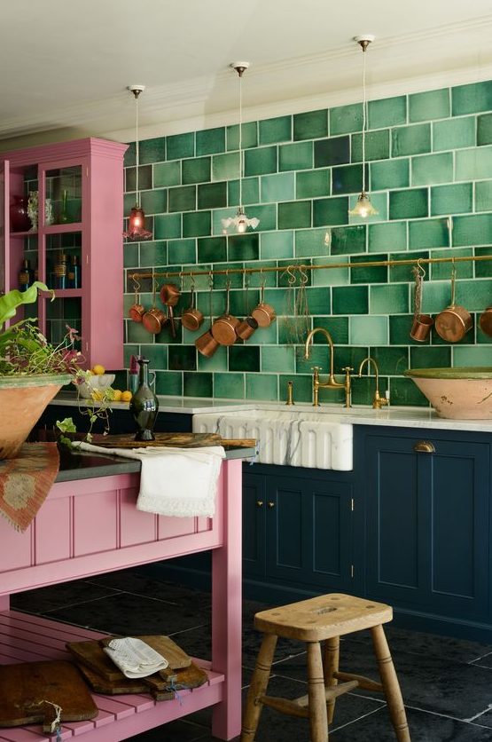 a bold maximalist kitchen with teal and pink cabinets, a green tile wall, a pink kitchen island and a wooden stool and brass cookware