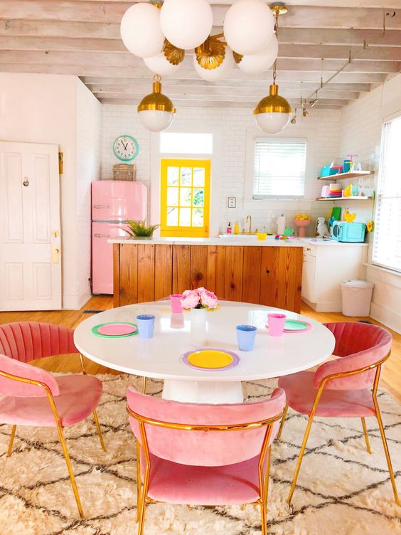 a candy-colored, maximalist kitchen with a neon yellow window, a pink refrigerator, pink velvet chairs, and pastel dishes
