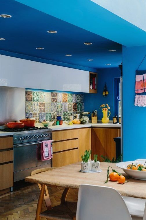 a colorful maximalist kitchen with bold blue walls and ceiling, white and stained cabinets, white countertops and a colorful backsplash