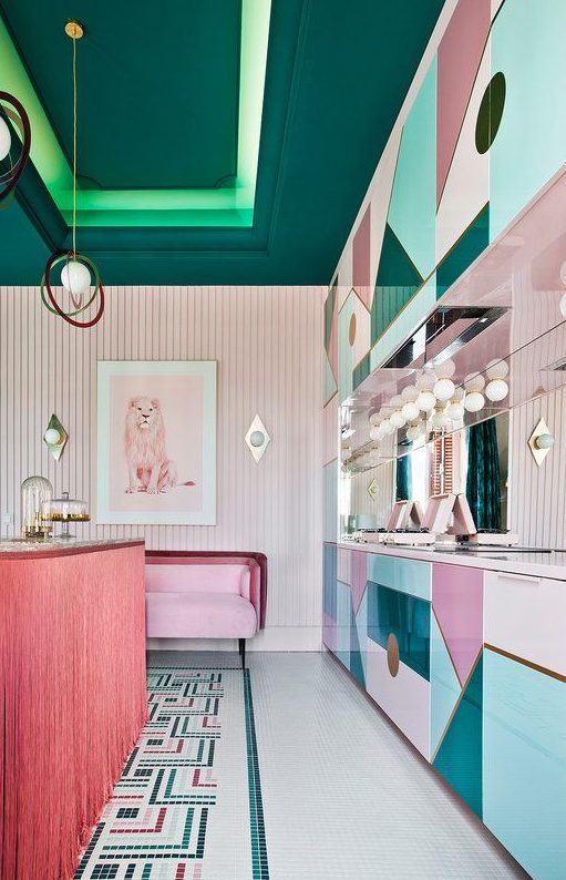 a sophisticated maximalist kitchen with pink, blush and blue geometric cabinets, a pink kitchen island, a pink sofa and a dark green blanket