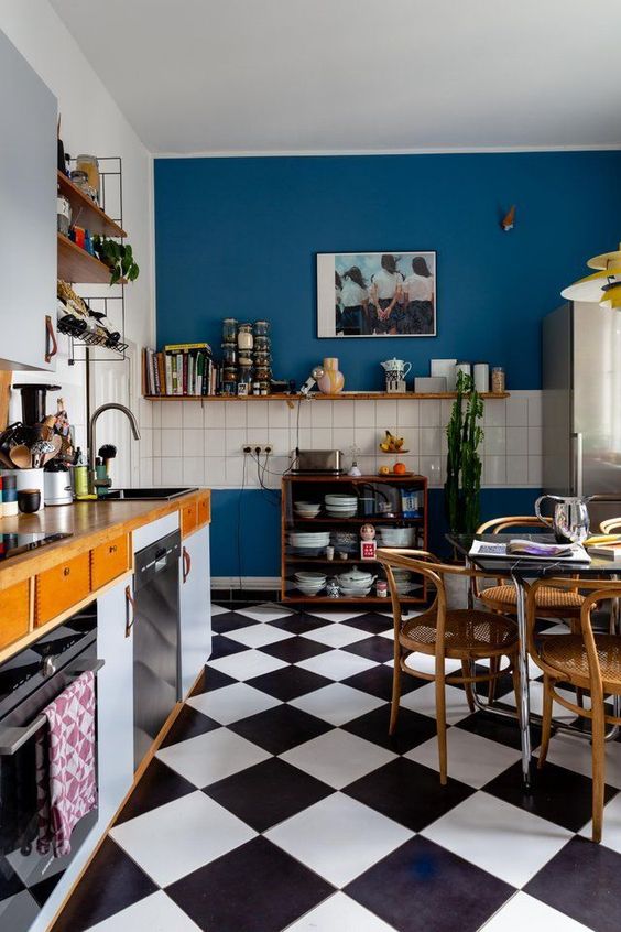 a stylish maximalist kitchen with bold blue walls, light blue and stained cabinets, a checkered floor and stained furniture