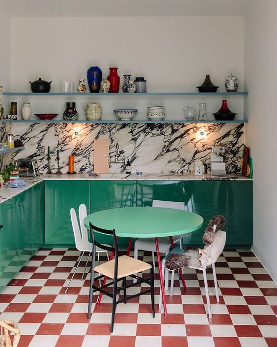 An elegant maximalist kitchen with emerald green cabinets, a red and white checkered floor, a white marble backsplash and countertop, and floating shelves with various decors