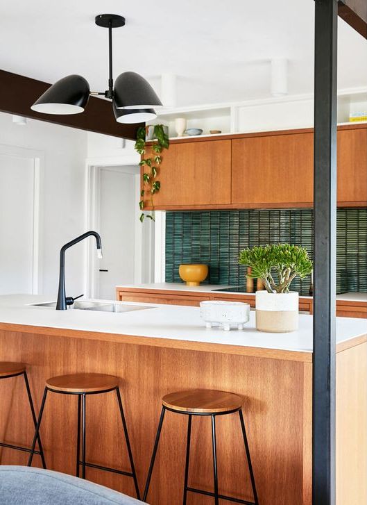 A stylish mid-century modern kitchen with light stained cabinets, white stone countertops, a green thin tile backsplash, black fixtures and potted plants