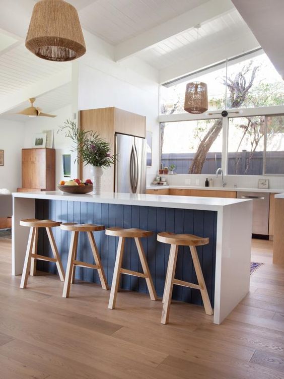 A mid-century modern kitchen with light stained cabinets, a white and navy island, white stone countertops, pendant lamps and views