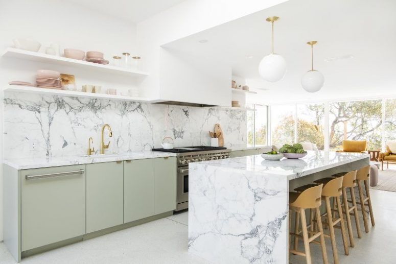 A glamorous mid-century modern kitchen with green cabinets, open shelving, a white range hood, shelves and a marble kitchen island