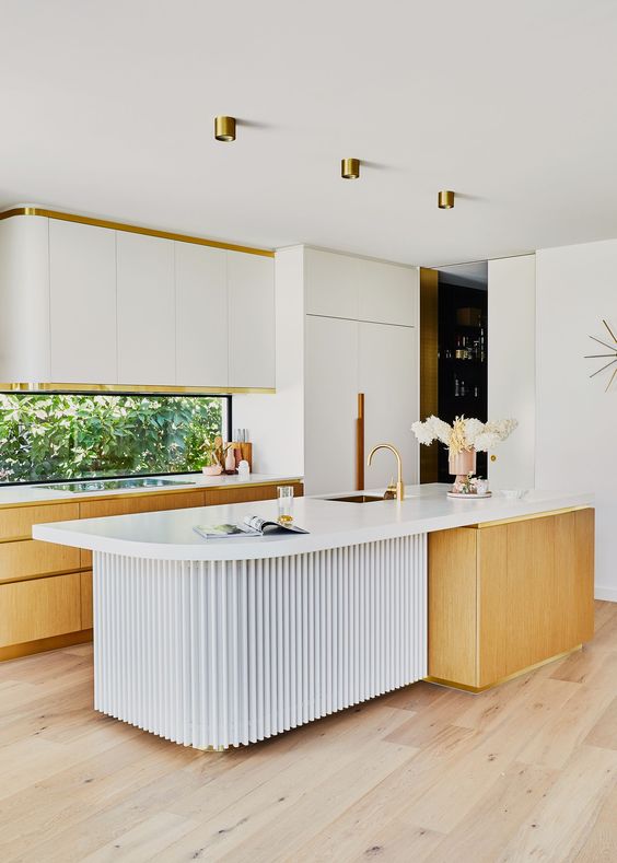 A sophisticated mid-century modern kitchen with white and light stained cabinets, a window backsplash, a sculptural island, and gold accents