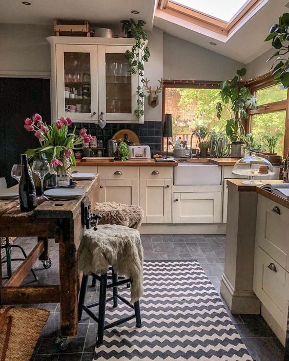 a cozy little kitchen with large windows and a skylight, vintage ivory furniture, black square tiles, a rough wooden table and lots of plants