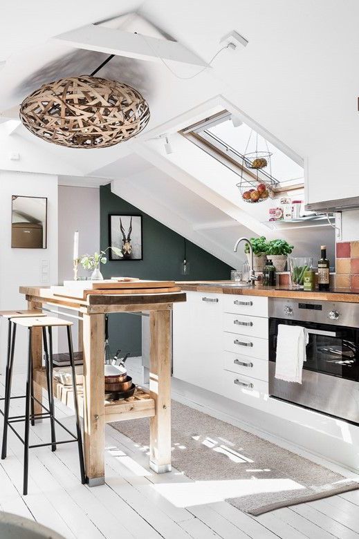 a beautiful attic kitchen with white cabinets, a tall wooden kitchen island and table, a beautiful wicker lamp and artwork