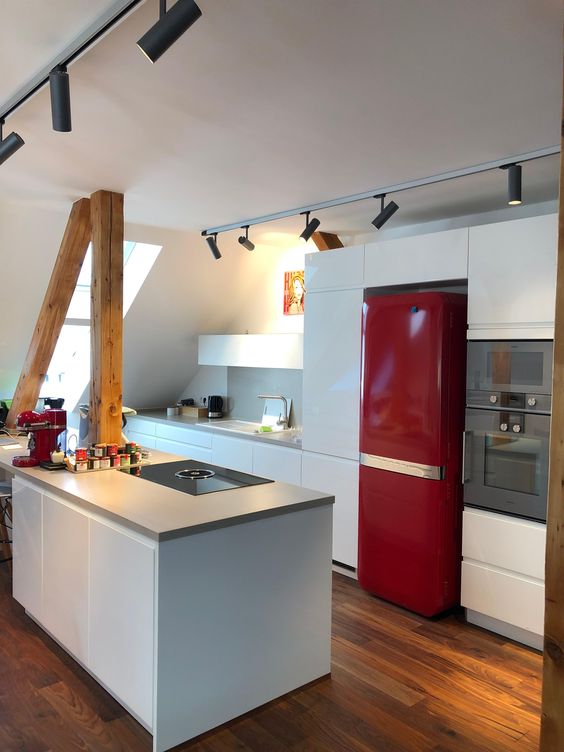 a minimalist attic kitchen with sleek white cabinets, a striking red refrigerator, wooden beams and lights across the room