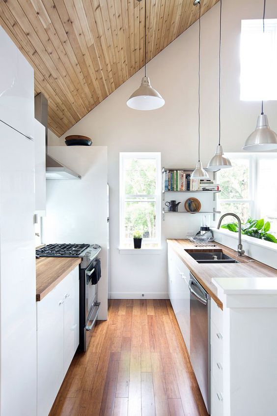 a small attic kitchen with a wood paneled ceiling, white cabinets and butcher block countertops, pendant lamps and lots of windows