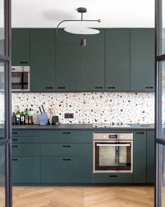 A chic teal kitchen with black countertops and a bright terrazzo backsplash and a statement chandelier is a very stylish space