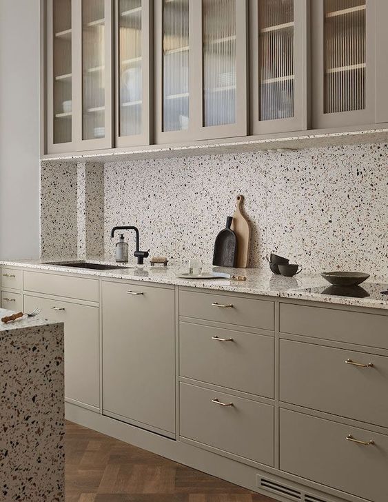 A gray kitchen with brass handles and striking white terrazzo countertops and backsplash and black fixtures is chic