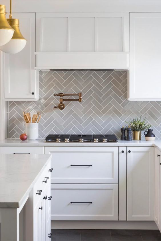 A chic white farmhouse kitchen with a gray herringbone backsplash and white countertops and gold and brass accents