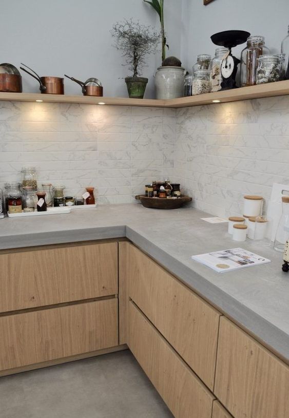 A stylish modern kitchen with light stained cabinets, concrete countertops, a wooden corner shelf with lights and a white marble tile backsplash
