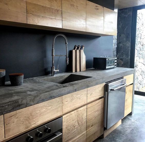 A stylish, minimalist kitchen with light-stained cabinets, a black backsplash, and a concrete countertop also has an industrial feel