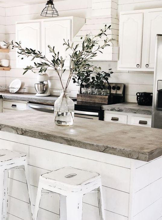 a white farmhouse kitchen with concrete countertops, a white plywood hood, and white shabby chic stools