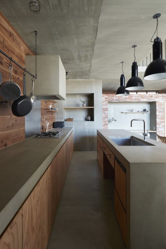 A light stained industrial kitchen with concrete countertops and hood, a brick wall and a light stained wall