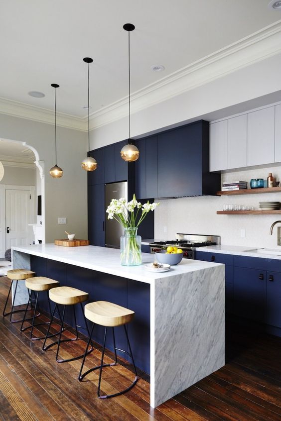 A beautiful navy and white kitchen with a white stone waterfall countertop and hanging lamps and wooden stools