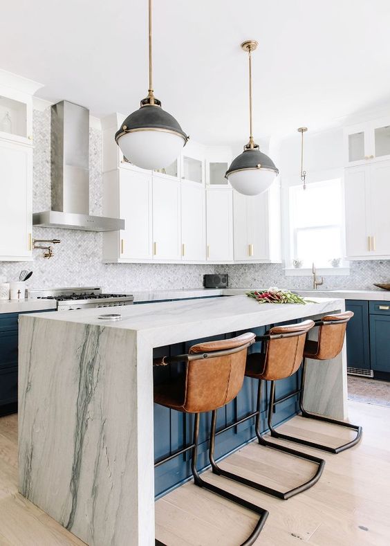 A white and navy kitchen with a navy island and white stone waterfall countertop and leather stools is pure chic