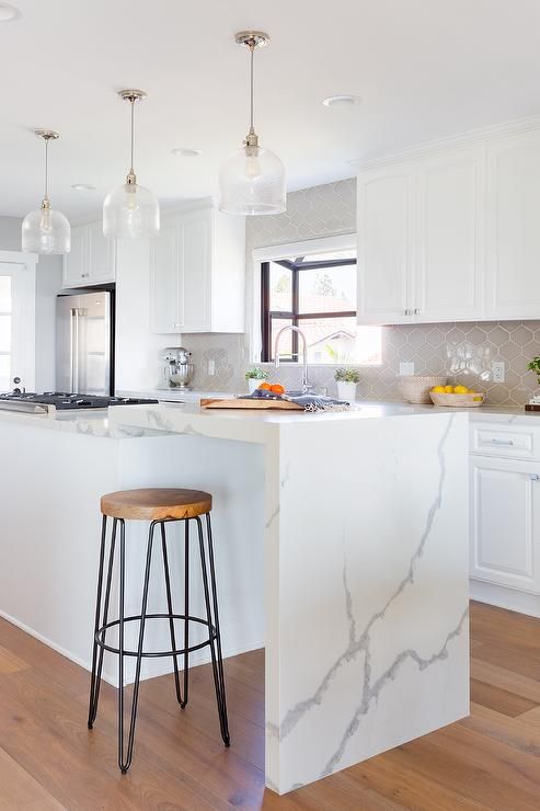 a sleek white kitchen with a gray tile backsplash and a kitchen island with a waterfall countertop as a breakfast bar