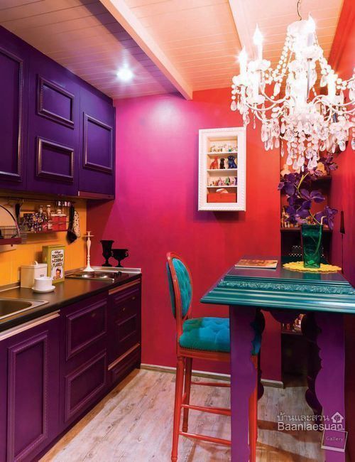 a colorful kitchen with pink and red walls, purple cabinets, black countertops and a yellow backsplash, a green table and a crystal chandelier