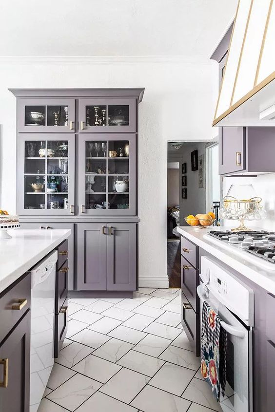 A pastel lavender kitchen with elegant vintage cabinets, white stone countertops, a stove and a vintage hood with gold edges