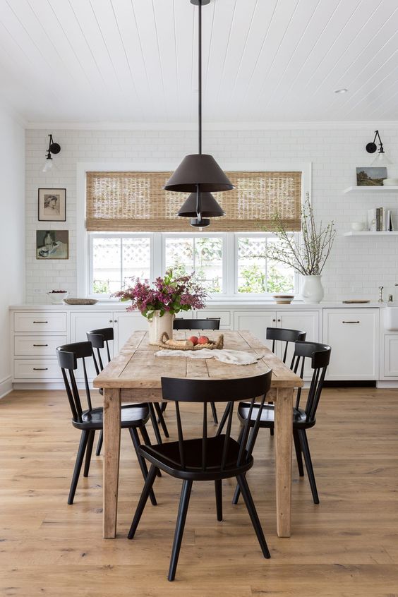 a cozy farmhouse kitchen with white base cabinets, a raw wood dining table, black chairs and black pendant lamps