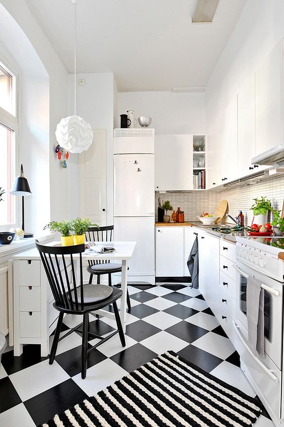 a small, eye-catching kitchen with white cabinets, a thin tile backsplash, a whimsical pendant lamp, a black and white floor, and a small dining area