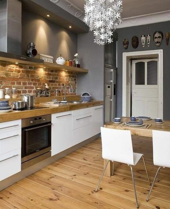 a stylish Scandinavian kitchen with white cabinets, wooden countertops, a brick backsplash, a floral chandelier, white chairs and a wooden table