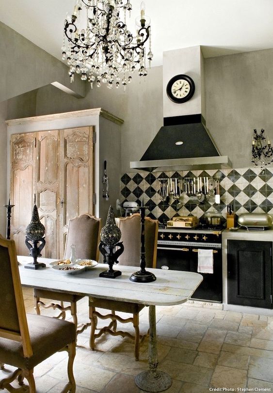a vintage kitchen with black and white cabinets, a crystal chandelier, a wooden table, vintage chairs and candle holders, and a modern clock