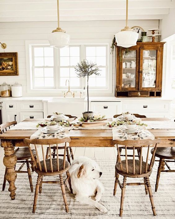 a white farmhouse kitchen with vintage wood cabinets, a vintage wood table, chairs and pendant lamps, and artwork