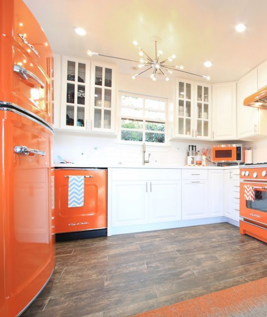 A striking, modern kitchen with white and orange cabinets, a sunlit chandelier, and an orange refrigerator is cool