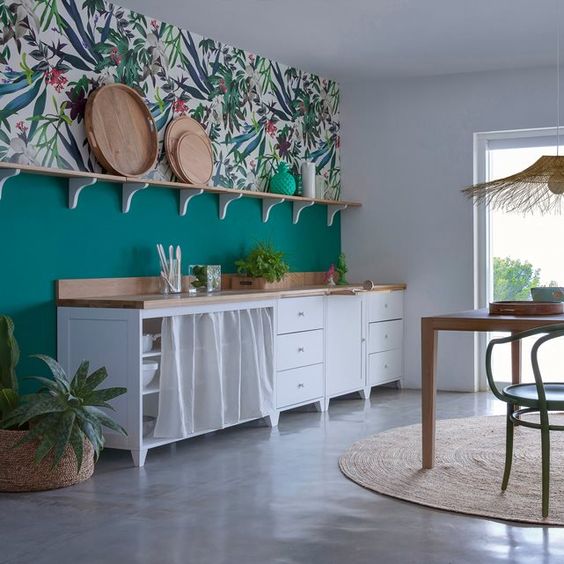 a bold tropical kitchen with an emerald green and light tropical wallpaper wall, white curtained cabinets and a wicker hanging lamp