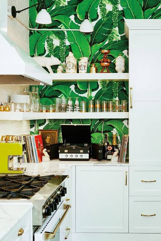 A glamorous tropical kitchen with banana leaf wallpaper, white cabinets, gold handles and wall sconces for a chic look