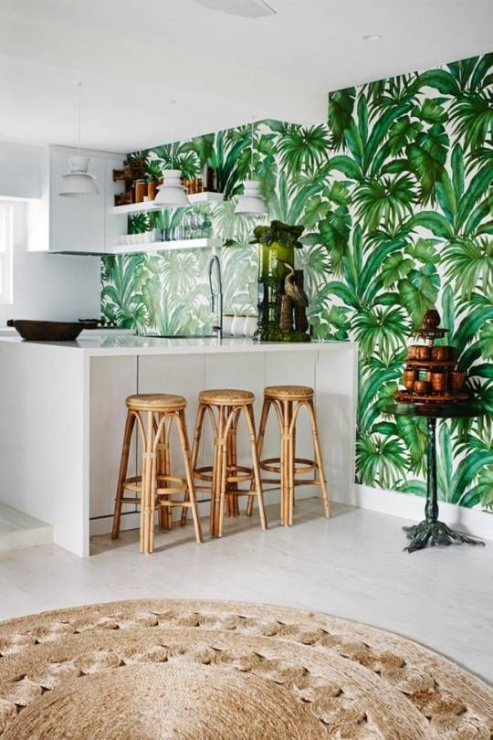 A stylish tropical kitchen with a wall of tropical leaves, sleek white cabinets, rattan stools and jute rugs is reminiscent of beaches