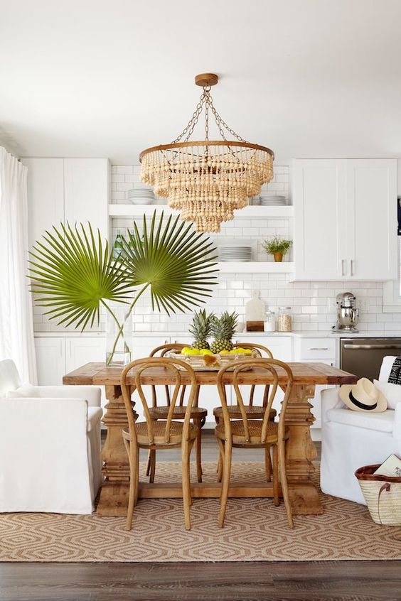 a tropical kitchen with white cabinets, a subway tile backsplash, a wooden table and chairs, and a beaded chandelier and tropical leaves