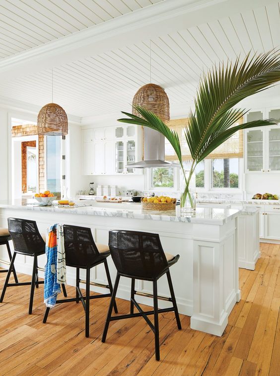 a white tropical kitchen with white cabinets and stone countertops, rattan pendant lamps and black stools