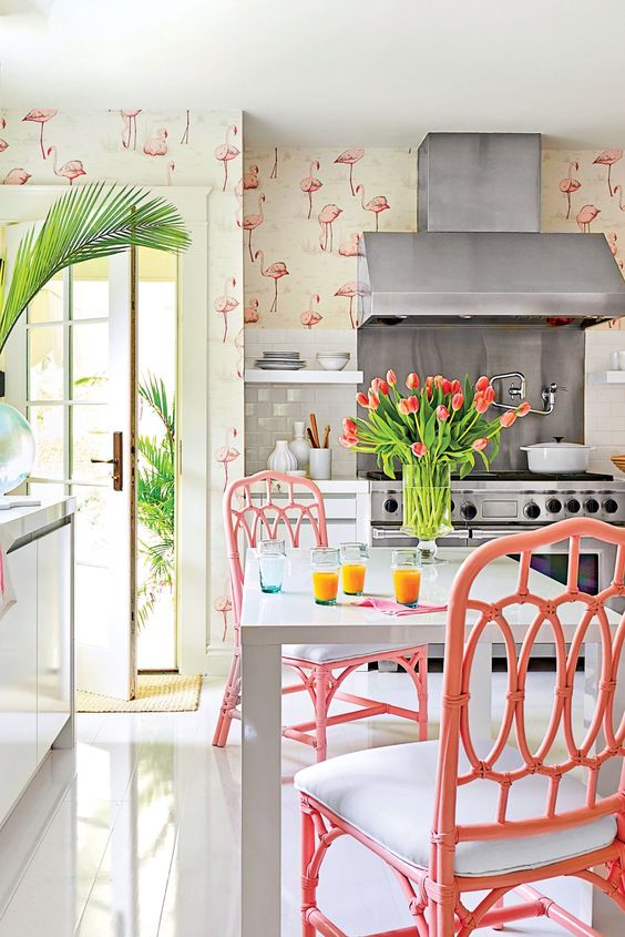 a whimsical tropical kitchen with flamingo wallpaper, white cabinets and a table, pink rattan chairs, and flowers and greenery