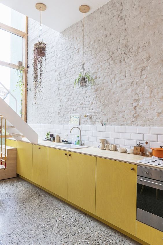 a modern kitchen with a brick wall and a white tile backsplash and yellow plywood cabinets