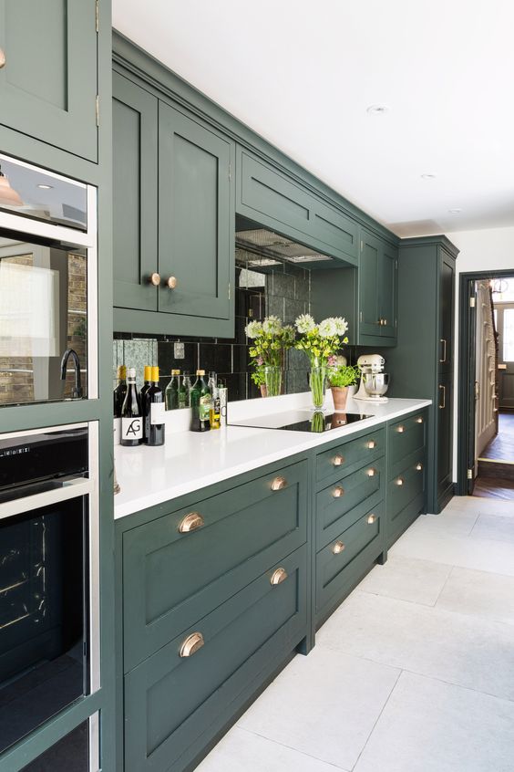 A sleek hunter green kitchen with white countertops, a mirrored tile backsplash, and brass accents