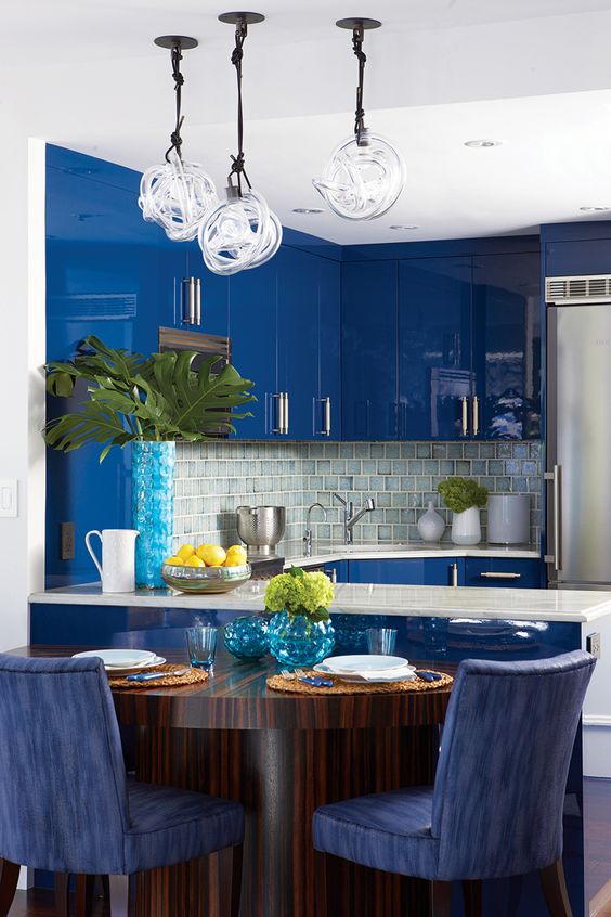 a bright blue kitchen with sleek and glossy cabinets, a neutral tile backsplash and hanging lamps, and white countertops