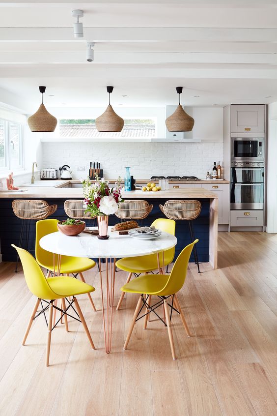 a cool tropical kitchen with a navy island as well as white cabinets, rattan stools and wicker lamps, bright yellow chairs and a round table