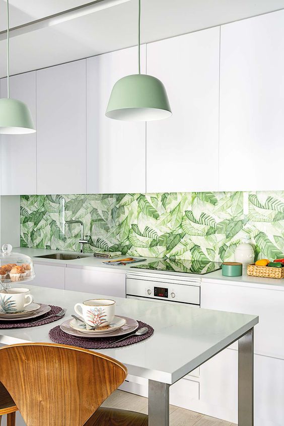 a modern tropical kitchen with sleek white cabinets, a tropical leaf patterned backsplash, green pendant lamps and wooden chairs