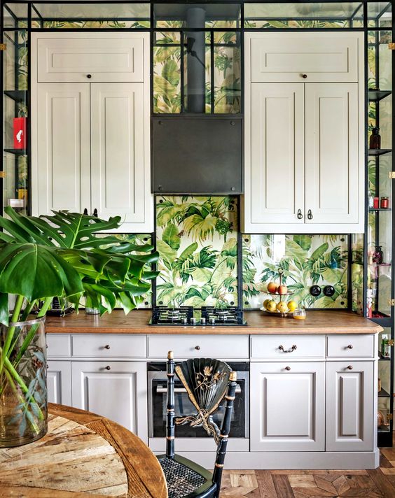 A vintage tropical kitchen with cream cabinets, a wooden table, black rattan chairs and tropical leaf wallpaper